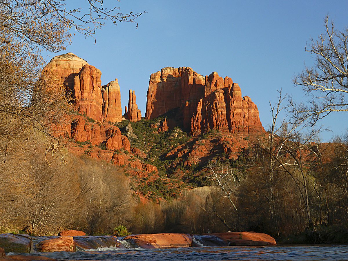 A red rock formation in the middle of a river with Cathedral Rock in the background.